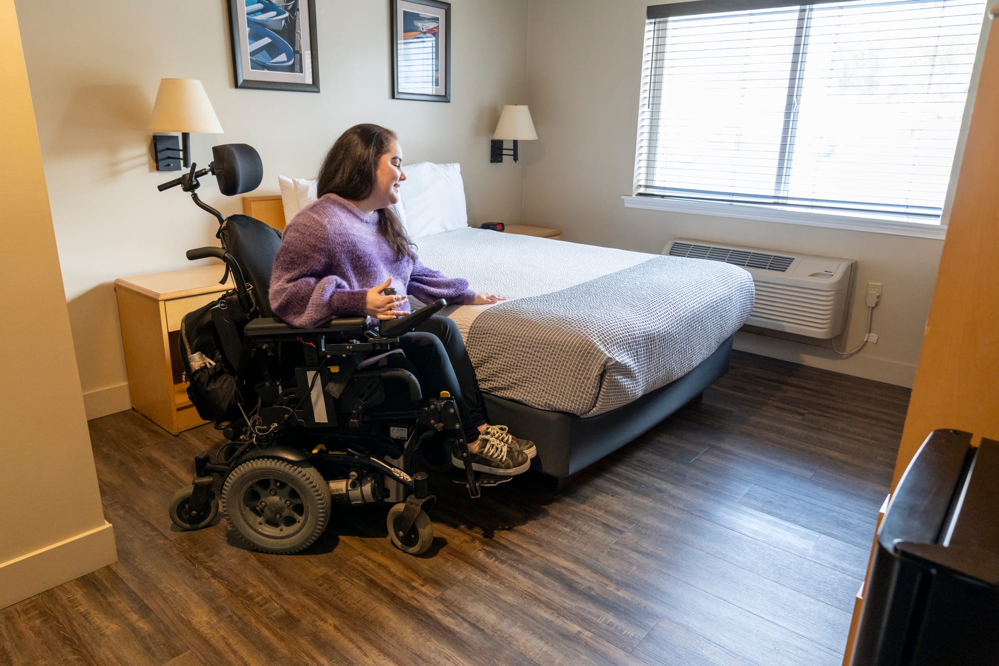 Power wheelchair-user next to an accessible bed in an accessible hotel room