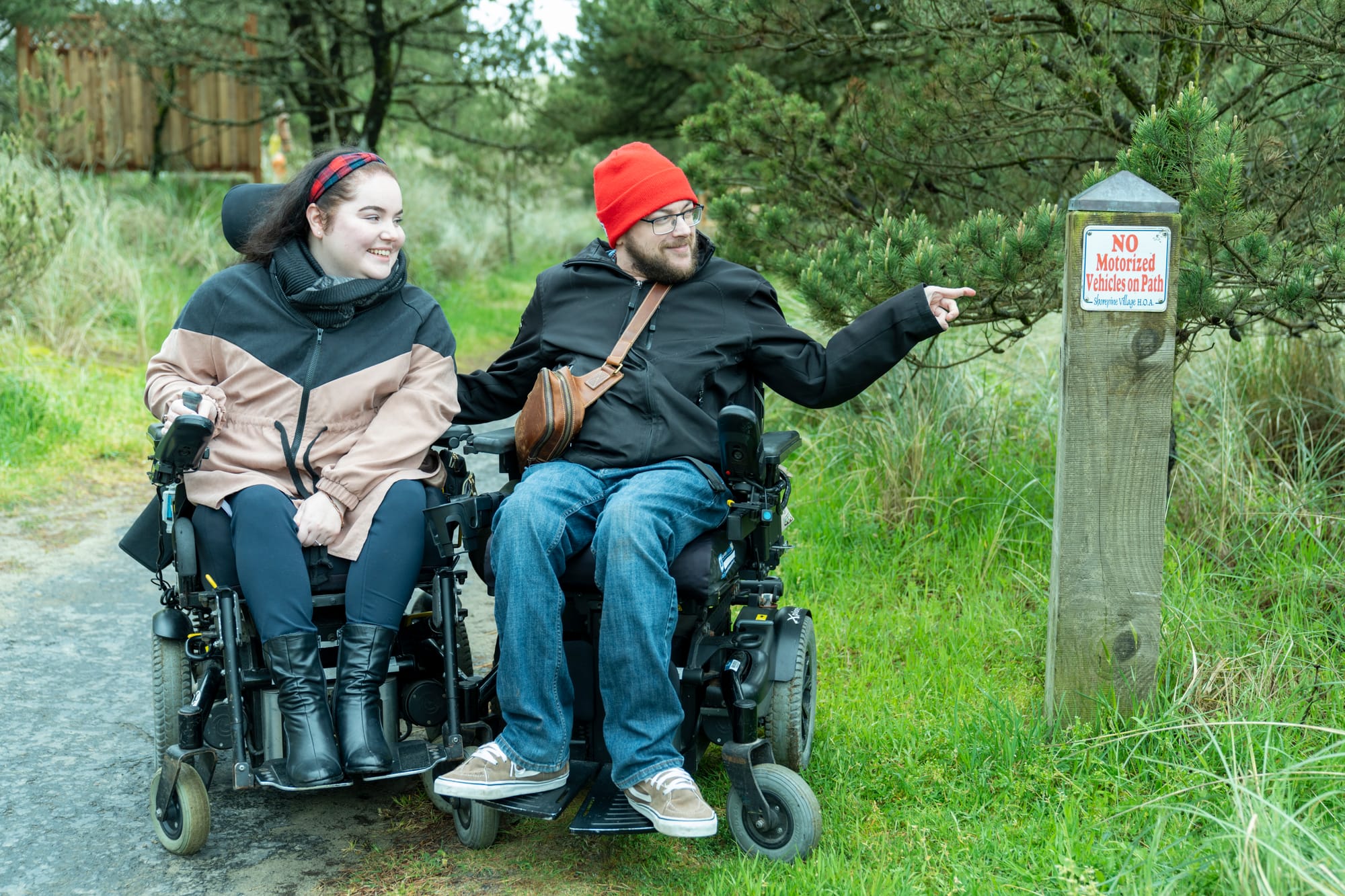 two power-wheelchair users on an accessible path with sign that reads "No Motorized Vehicles on Path"