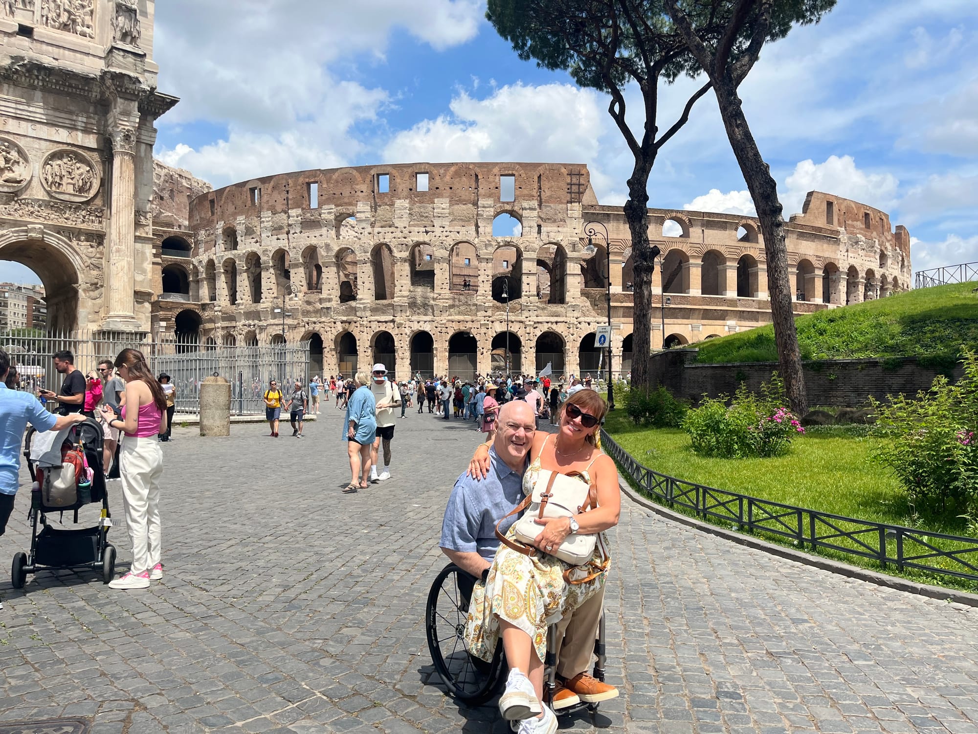 Wife sitting on husbands lap, who is sitting in a wheelchair, with Colosseum in the background
