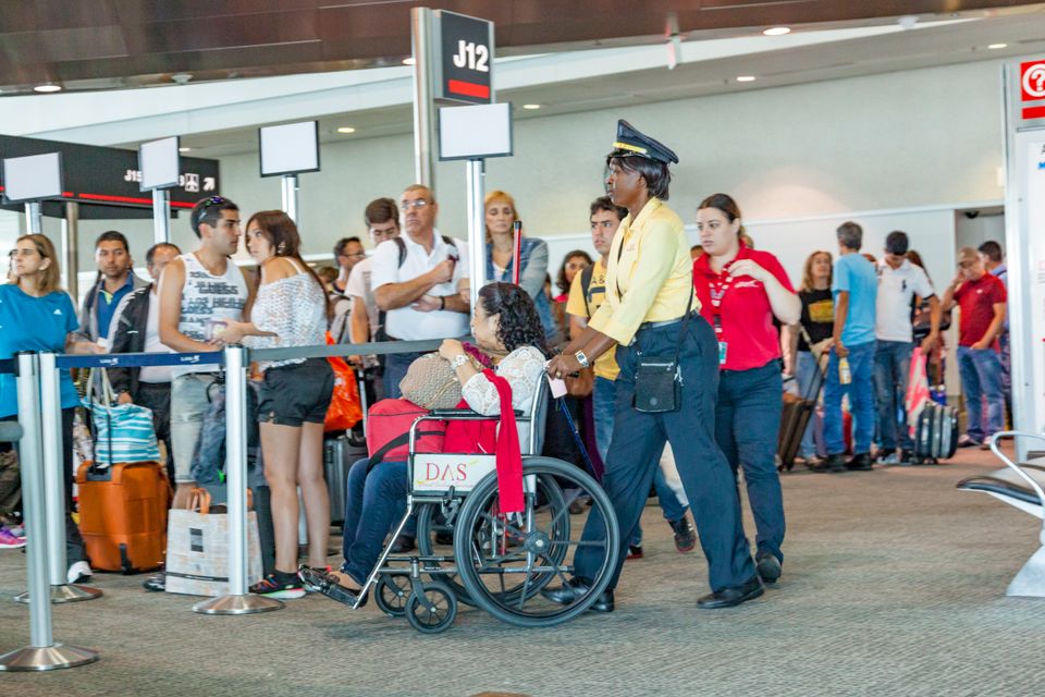 Wheelchair-user carrying luggage on her lap while wheelchair attendant assists her through the airport.