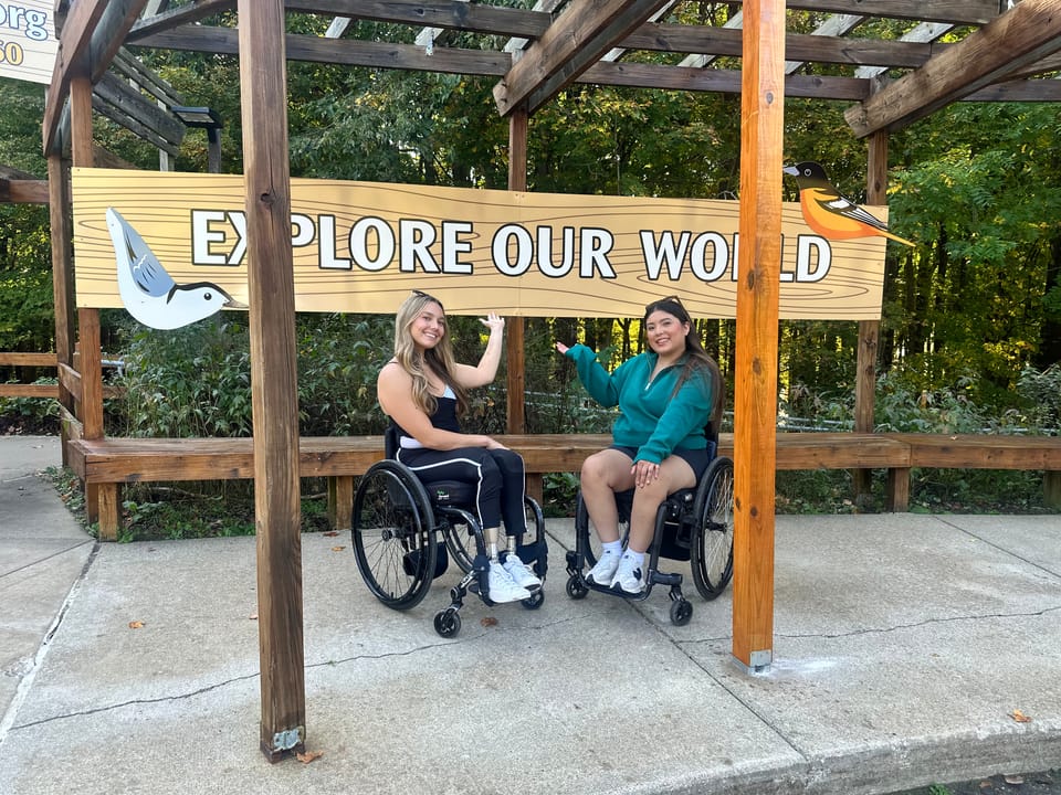 Two wheelchair users at Kalamazoo Nature Center in front of sign that says "Explore Our World"